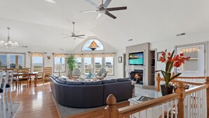 A spacious living area with a large dark-colored sectional sofa, a dining area with wooden chairs, a fireplace, and a TV mounted on the wall. - Overdraft: Steps to Beach, Pool, Hot Tub, 7BR OBX (Southern Shores)