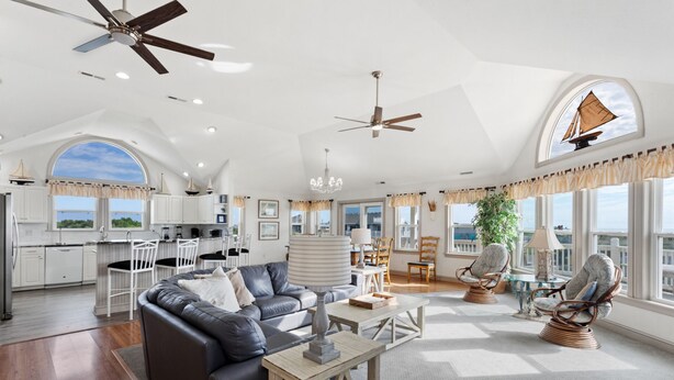 A spacious living area with a dark leather sofa, wooden coffee table, and wicker chairs. There are ceiling fans, a chandelier, and large windows offering views of the outdoors. - Overdraft: Steps to Beach, Pool, Hot Tub, 7BR OBX (Southern Shores)