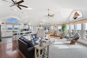 A spacious living area with a dark leather sofa, wooden coffee table, and wicker chairs. There are ceiling fans, a chandelier, and large windows offering views of the outdoors.