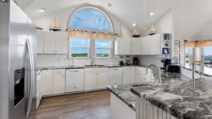 A modern kitchen with white cabinets, a granite countertop, and a large window with a view of the outdoors. - Overdraft: Steps to Beach, Pool, Hot Tub, 7BR OBX (Southern Shores)