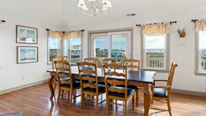 A dining room with a wooden table and chairs, large windows with curtains, and a chandelier. - Overdraft: Steps to Beach, Pool, Hot Tub, 7BR OBX (Southern Shores)