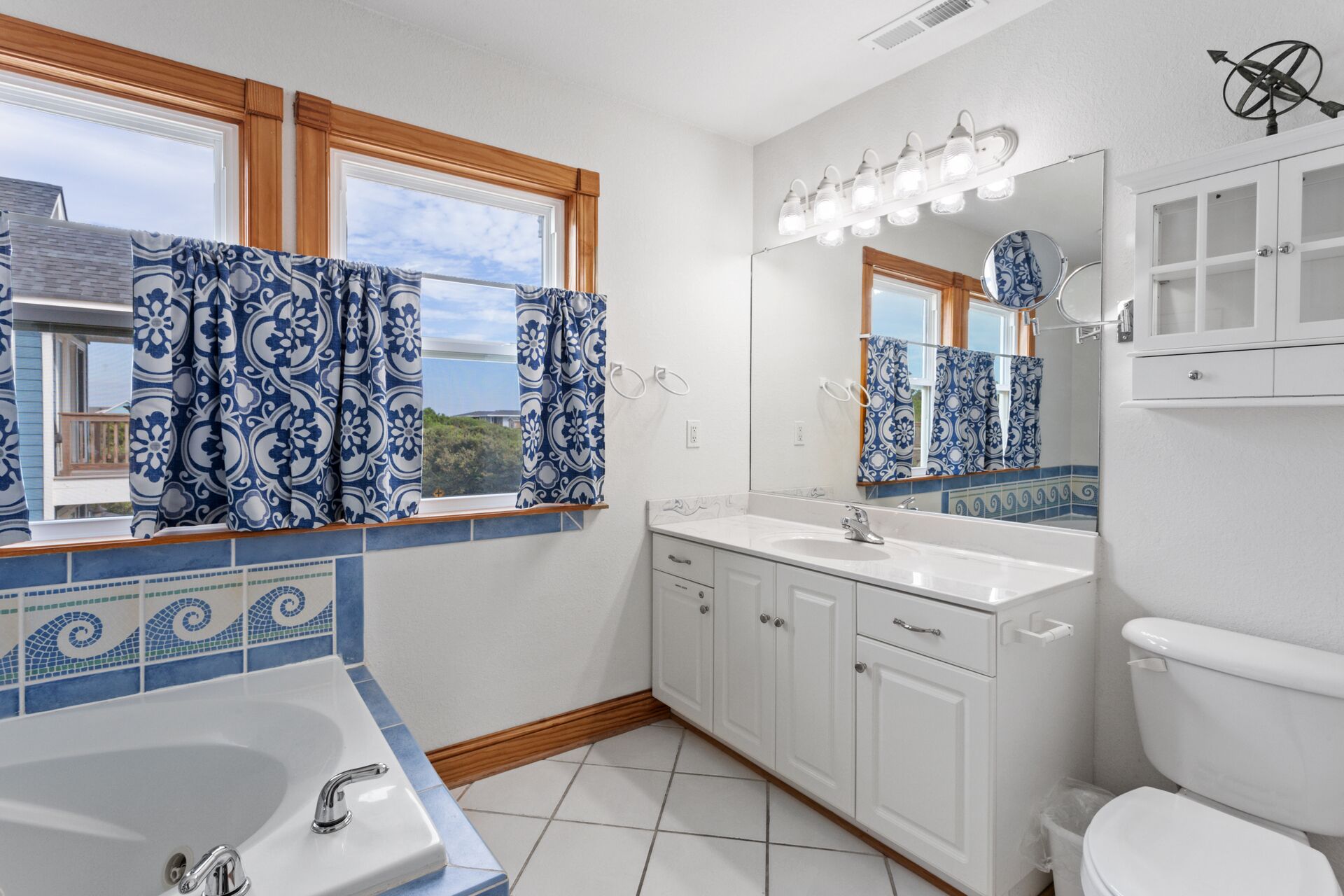 A bathroom with a bathtub, sink, and toilet. There are windows with curtains, a mirror with lights, and a tiled backsplash.