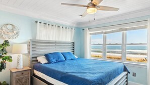 A bedroom with a bed, a nightstand, a lamp, a ceiling fan, and a window with a beach view.