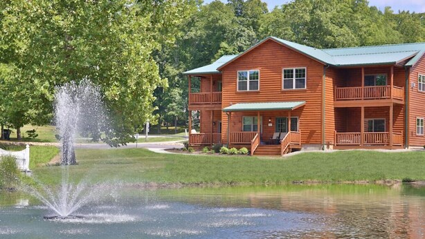 A two-story log cabin with a porch and a pond with a fountain.