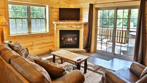 A cozy log cabin interior with a fireplace, a television, a sofa, and a patio area with outdoor furniture.