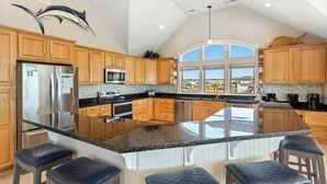 A spacious kitchen with wooden cabinets, a large island with bar stools, stainless steel appliances, and a view through large windows. - Just Fab * Oceanfront * Elevator * Saltwater Pool * Hot Tub * Game Room * Views (Nags Head)