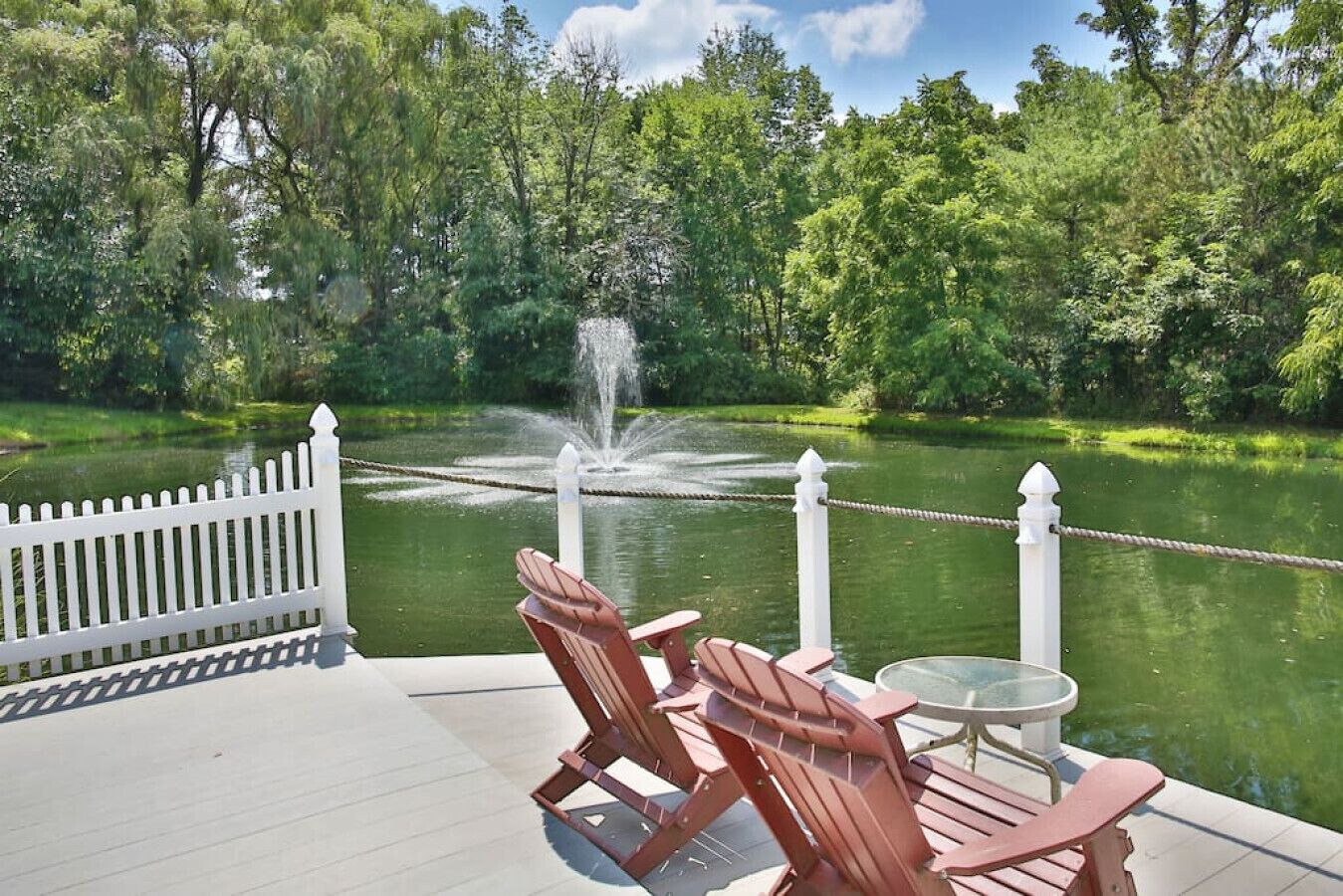 Um terraço com cadeiras Adirondack e uma mesa, com vista para um lago com uma fonte.