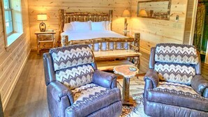 Une chambre aménagée dans un chalet en rondins, avec un grand lit, deux fauteuils en cuir, une petite table et une fenêtre.