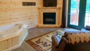 L’intérieur d’une cabane en rondins, avec un plancher en bois, une cheminée, un téléviseur fixé au-dessus, une baignoire et une porte-fenêtre coulissante donnant sur l’extérieur.