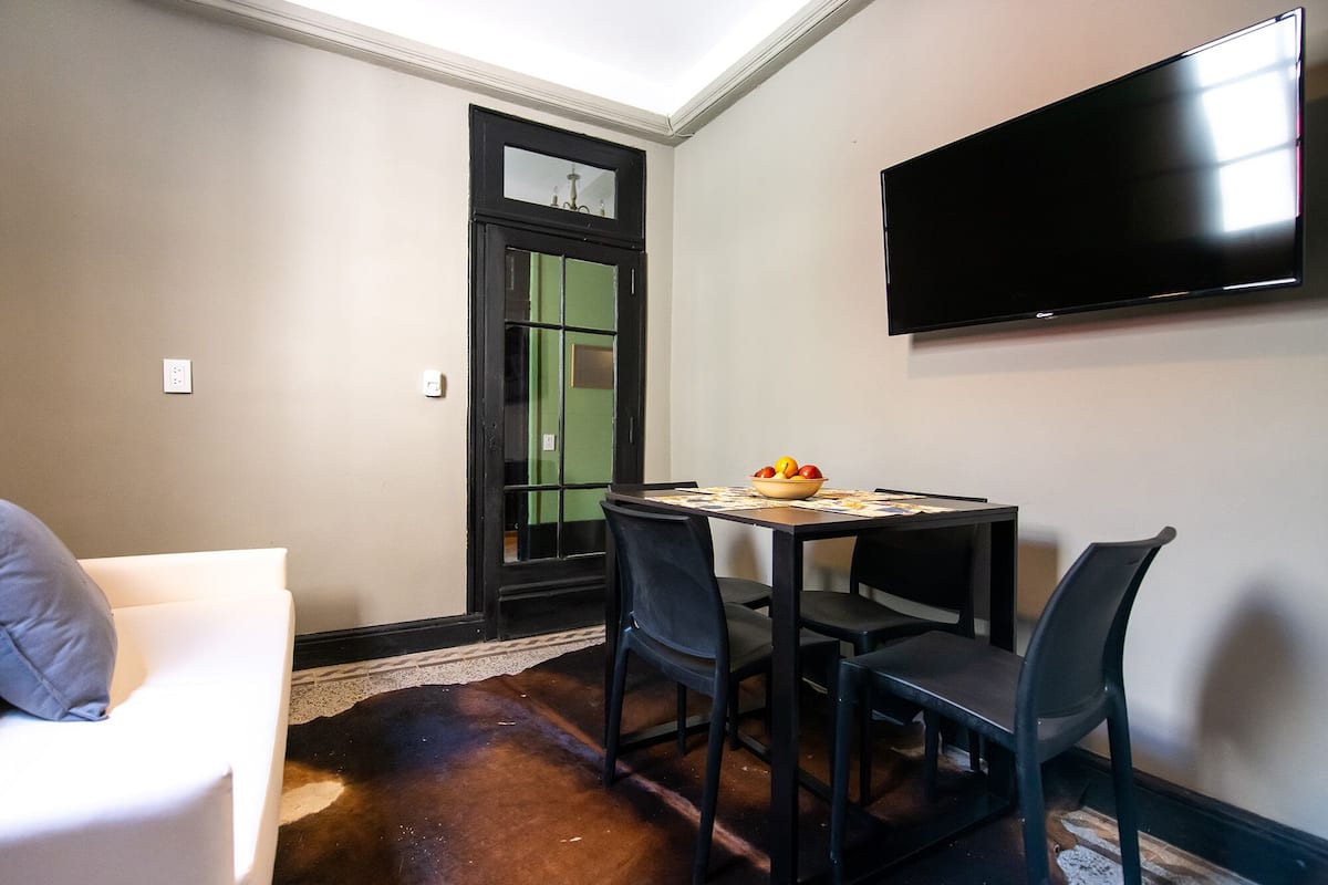 A small dining area with a table, chairs, a bowl of fruit, a flat-screen TV mounted on the wall, and a door with a green frame.