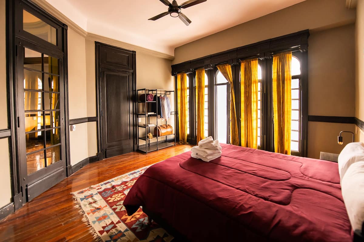 A hotel room with a large bed, a ceiling fan, a wooden floor, and a window with curtains.