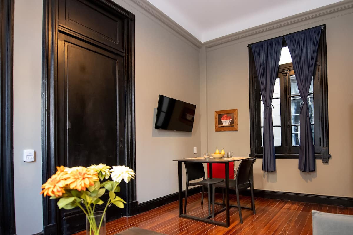 A room with a wooden floor, a dining table with chairs, a TV mounted on the wall, and a window with curtains.