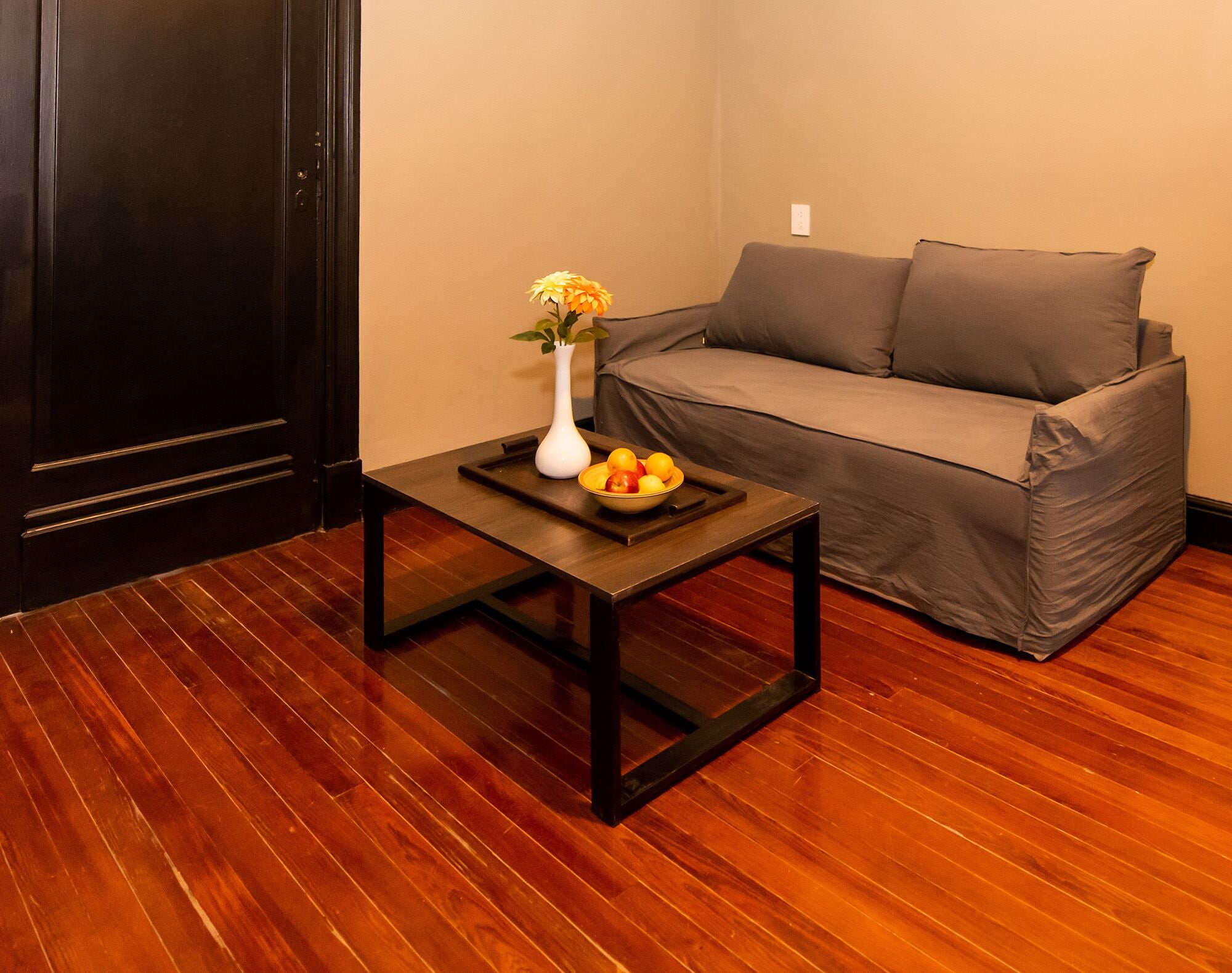 Um quarto com piso de madeira, uma mesa preta com uma tigela de frutas e um vaso, e um sofá cinza.
