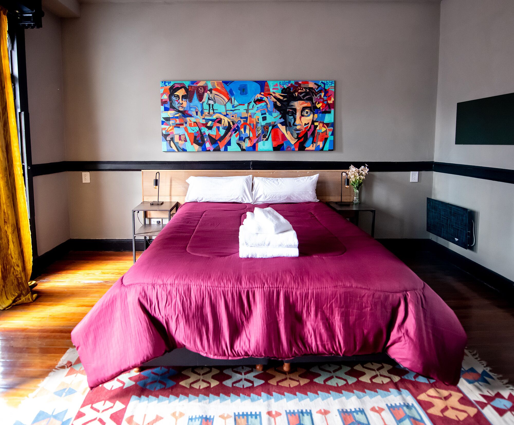 A hotel room with a large bed, a painting on the wall, a bedside table with a vase of flowers, and a patterned rug.