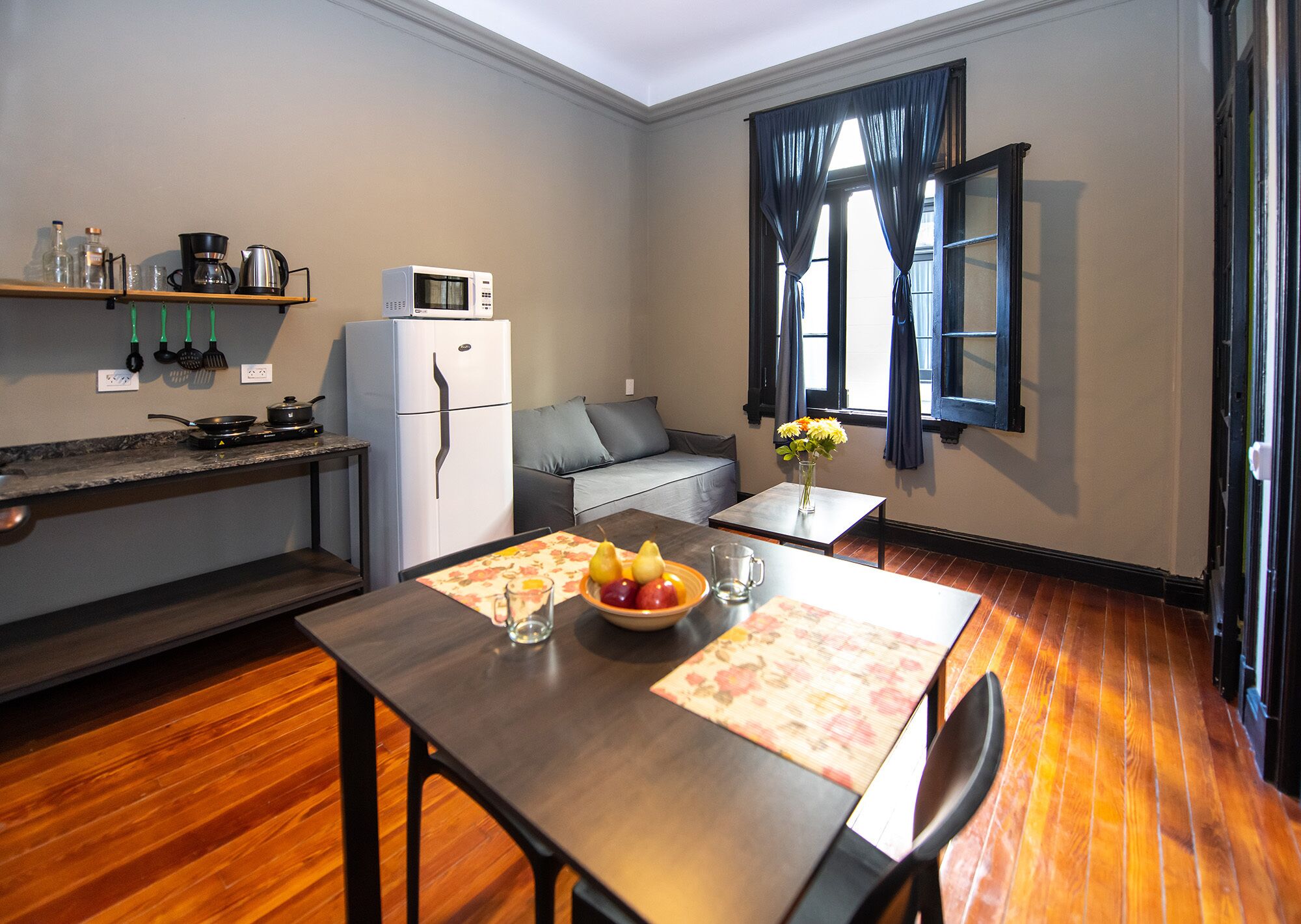 Cocina funcional con refrigerador, microondas y estufa. Hay una mesa de comedor con un frutero y un florero. Un sofá está apoyado contra la pared, y se ve una ventana con cortinas.
