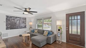 Un salon moderne avec un canapé d’angle, un ventilateur de plafond, une grande œuvre d’art représentant un paysage urbain et une porte dotée de panneaux en verre dépoli.