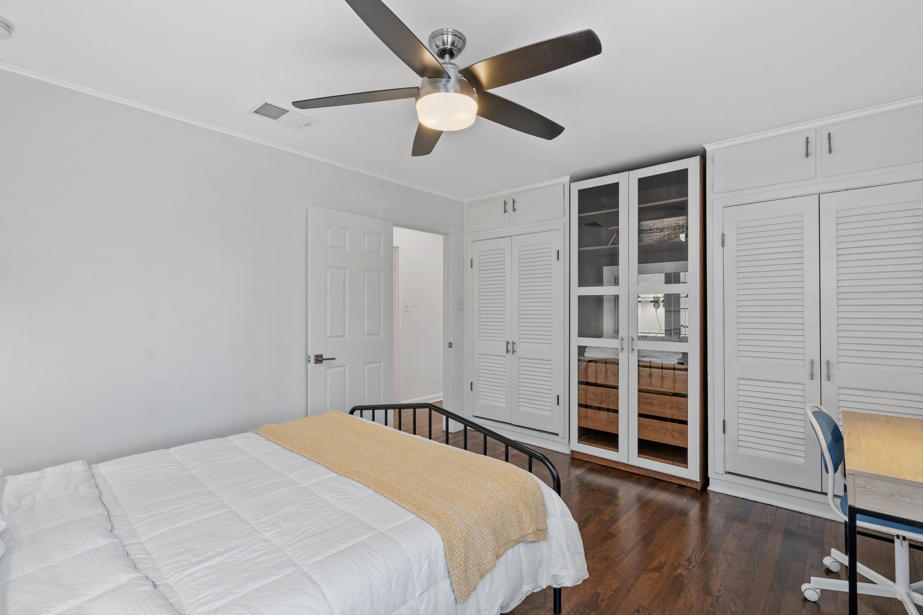 Une chambre à coucher avec un lit, un ventilateur de plafond, des armoires blanches et un bureau en bois.