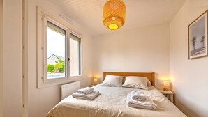 Una cama bien hecha con sábanas y toallas blancas, un cabecero de madera, lámparas de noche y una ventana con vistas a zonas verdes.