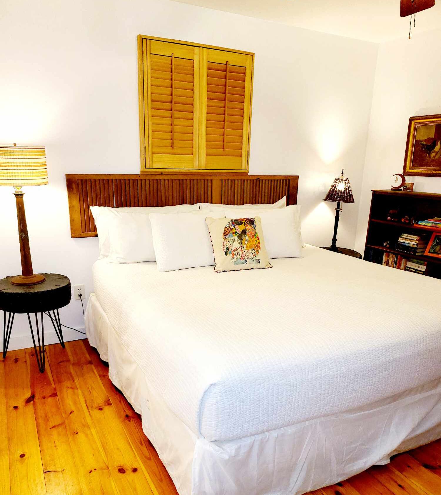 Una cama bien hecha con sábanas blancas, cabecera de madera y una ventana con contraventanas. Junto a la cama, una pequeña mesa redonda con una lámpara y una estantería con libros y un objeto decorativo a la derecha.