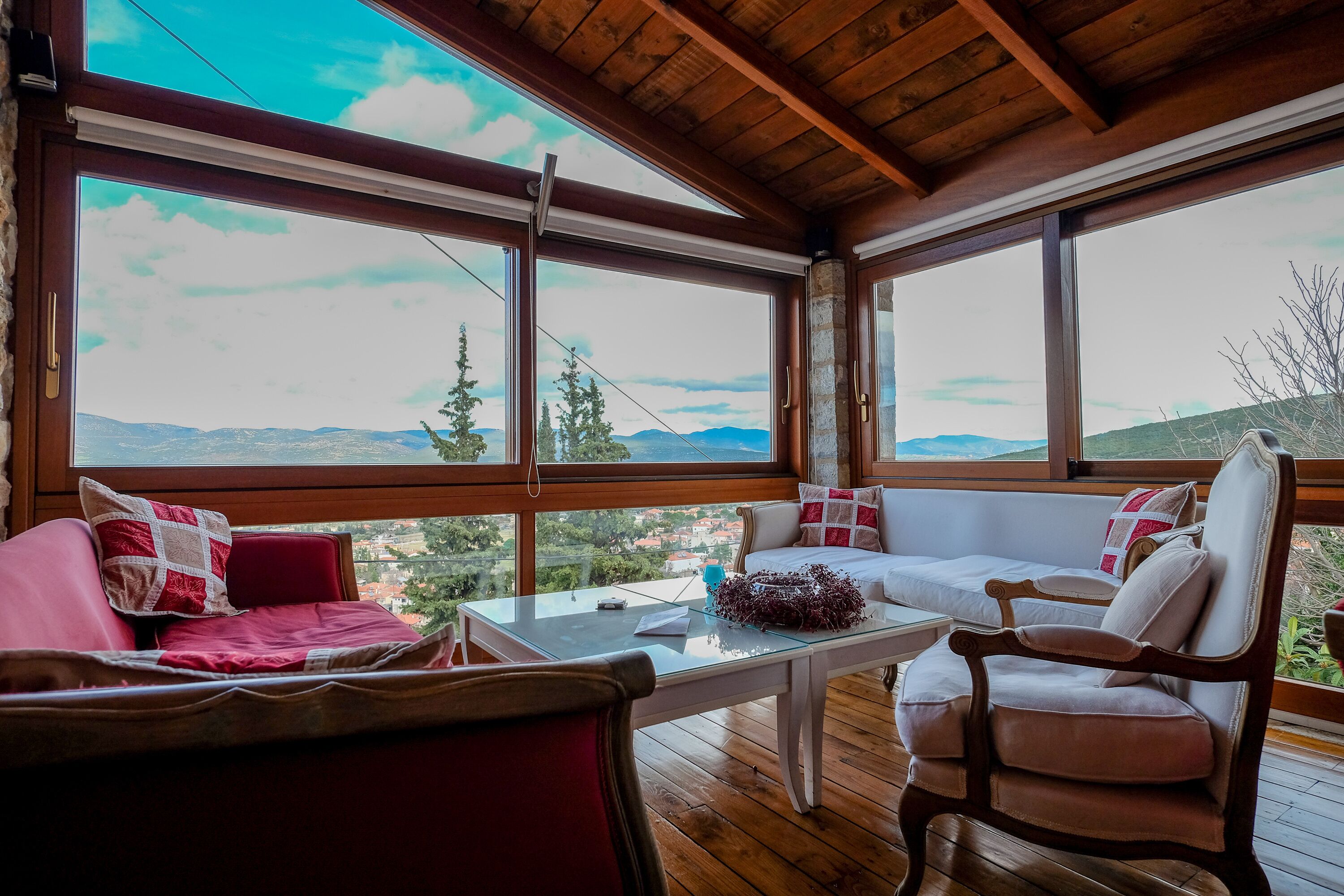 A cozy room with a wooden ceiling, large windows, a red sofa, a white sofa, and a wooden chair.