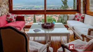 A cozy room with a large window offering a view of a town and mountains. There is a white table with a decorative bowl, red and white cushions, and wooden chairs. - KIRIAKI GUESTHOUSE & SUITES (Amfikleia-Elateia)