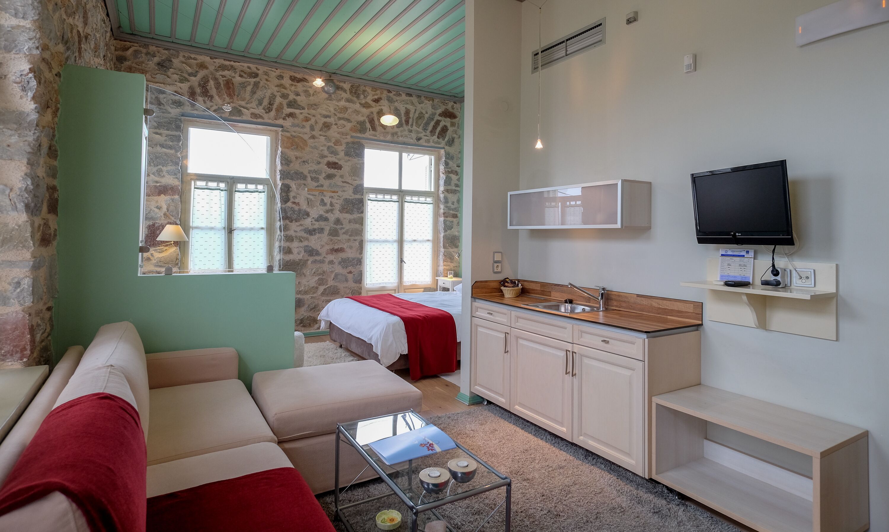 A room with a stone wall, a bed, a sofa, a small kitchen area, and a television.