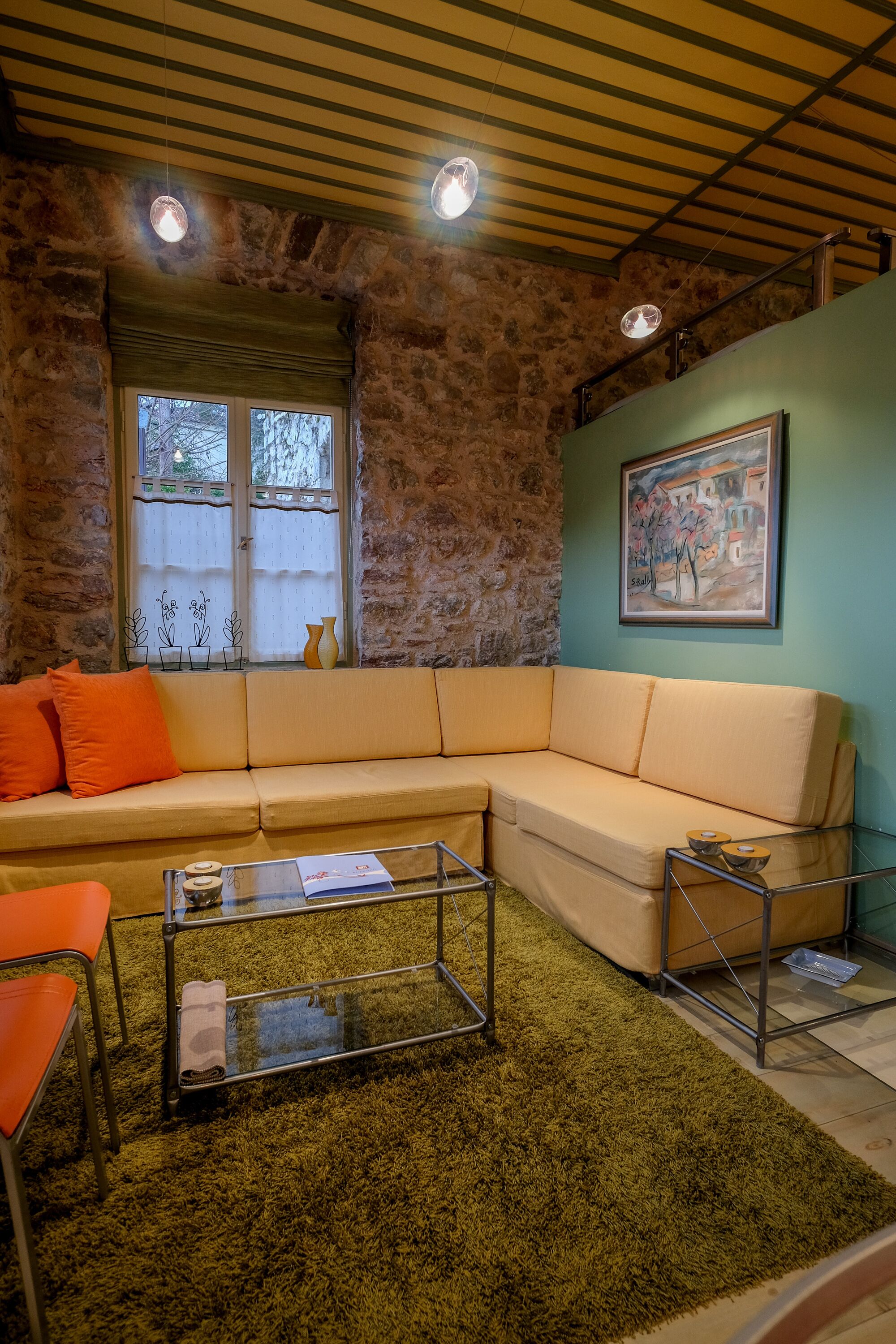 A cozy living room with a beige sectional sofa, orange cushions, a glass coffee table, and a green shag rug. A window with a view of trees is visible, and a painting hangs on the wall.
