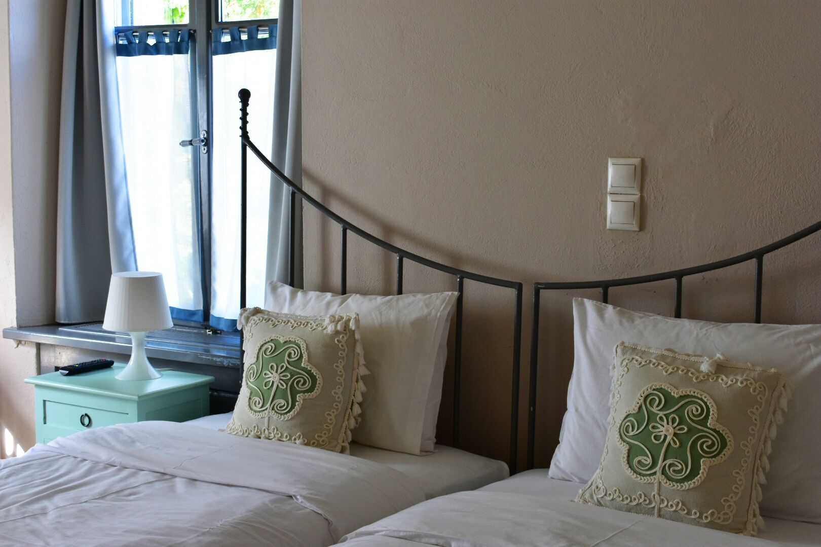 Quarto com cama, duas almofadas estampadas com flores, mesa de cabeceira com abajur e janela com cortinas.