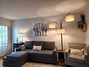Ein modernes Wohnzimmer mit einem grauen Ecksofa, einem kleinen Beistelltisch, einer Stehlampe und einem dekorativen Bild an der Wand.