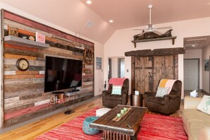 A living room with a rustic wooden wall, a flat-screen TV, two brown armchairs, a coffee table, and a red rug. - Casa Copano by Miss Kitty’s (Rockport)