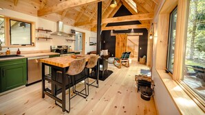 Interior rústico de uma cabana, com área de jantar de madeira, cozinha e janelas amplas com vista para uma floresta.