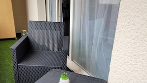 En terrasse med en mørk fletsofa og tilhørende skammel, en lille hvid plantekasse med en grøn plante, og en glasdør ind til et indendørs rum.