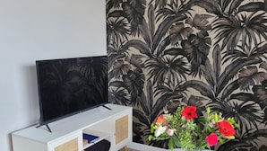 En moderne stue med et fladskærms-TV på et hvidt skab, et lille hvidt bord med en vase blomster, og tapet med et stort blomstermønster.