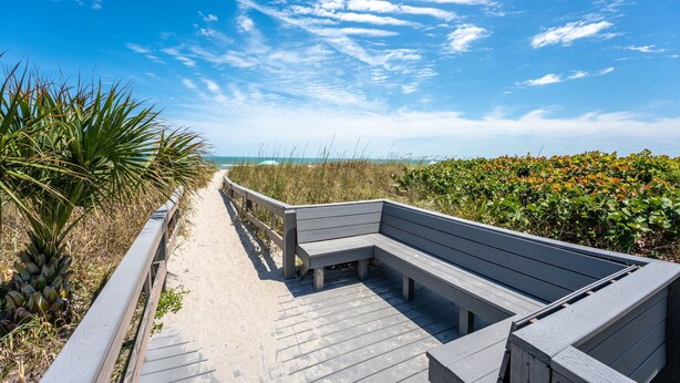 Une promenade en bois, agrémentée de bancs, qui mène à une plage bordée de dunes et de végétation.