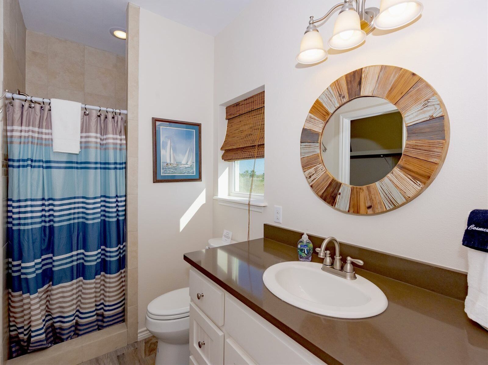 Un baño con cortina de ducha a rayas, un cuadro enmarcado con barcos de vela, un espejo redondo con diseño de veta de madera y un lavabo blanco con encimera de madera.