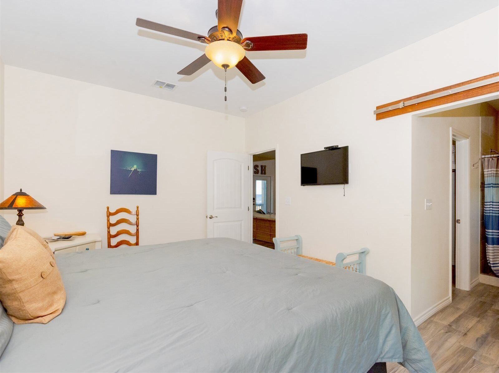 Un dormitorio con una cama, ventilador de techo, televisión, puerta que da a un baño y un cuadro en la pared.