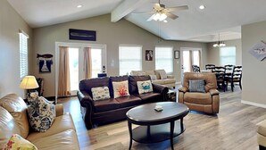 A spacious living room with a brown leather sofa, a round coffee table, and a tan armchair. There is a dining area with a table and chairs in the background. - A Fin N Feather by Miss Kitty’s (Rockport)