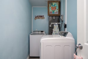 Un cuarto de lavandería con lavadora y secadora, un letrero que dice “Laundry Everything Comes Out and the Wash”, y un cuadro colorido sobre la secadora.