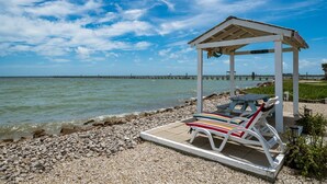A beachside gazebo with a table and chairs, overlooking a calm sea and a pier in the distance.