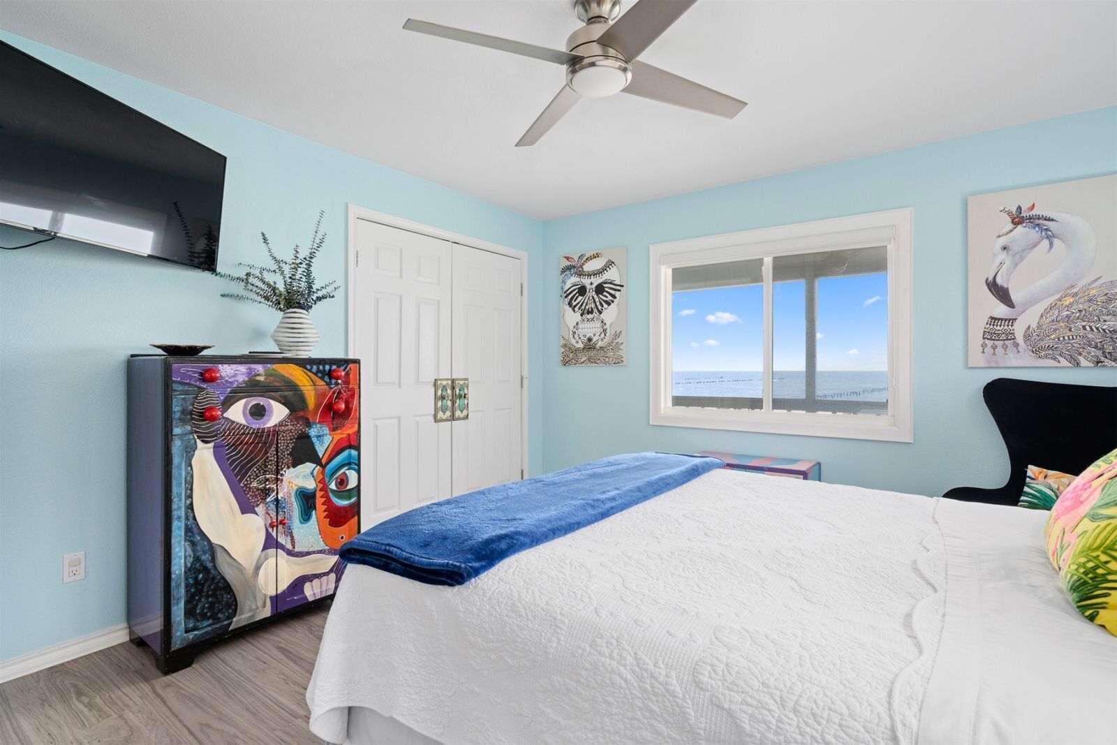 Un dormitorio con cama, ventilador de techo, ventana con vistas, puerta y televisor montado en la pared.