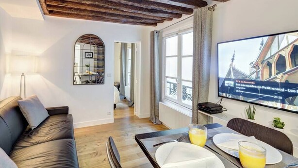 A modern living room with a wooden ceiling, a sofa, a dining table with plates and glasses, and a TV displaying a news channel. - Chic Marais Retreat | Steps to Cafes & Notre Dame (Paris)