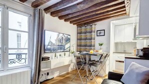 A compact living space with a dining area, kitchenette, and a large window with curtains. - Chic Marais Retreat | Steps to Cafes & Notre Dame (Paris)