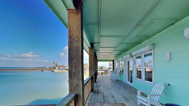 A wooden deck with a railing, a covered area with a view of the water, and a small table. - Pelican Point by Miss Kitty’s (Rockport)
