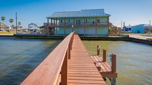 A wooden pier leading to a two-story building with a blue roof, situated by a body of water. - Pelican Point by Miss Kitty’s (Rockport)