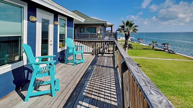 A wooden deck with two turquoise chairs, a view of the ocean, and a building with a balcony. - Fisherman Luv by Miss Kitty’s (Rockport)