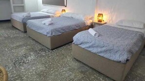 Ein Hotelzimmer mit drei Einzelbetten, jedes mit einer gemusterten Tagesdecke und einem gerollten Handtuch. An jeder Seite der Betten befindet sich ein Nachttisch mit einer Lampe.