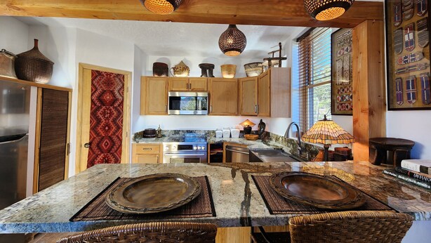 A modern kitchen with wooden cabinets, a stainless steel refrigerator, a microwave, and a stove. There are hanging pendant lights and a woven basket on the counter. - Come experience a taste of Africa nestled in the foothills of Santa Fe.  (Santa Fe)