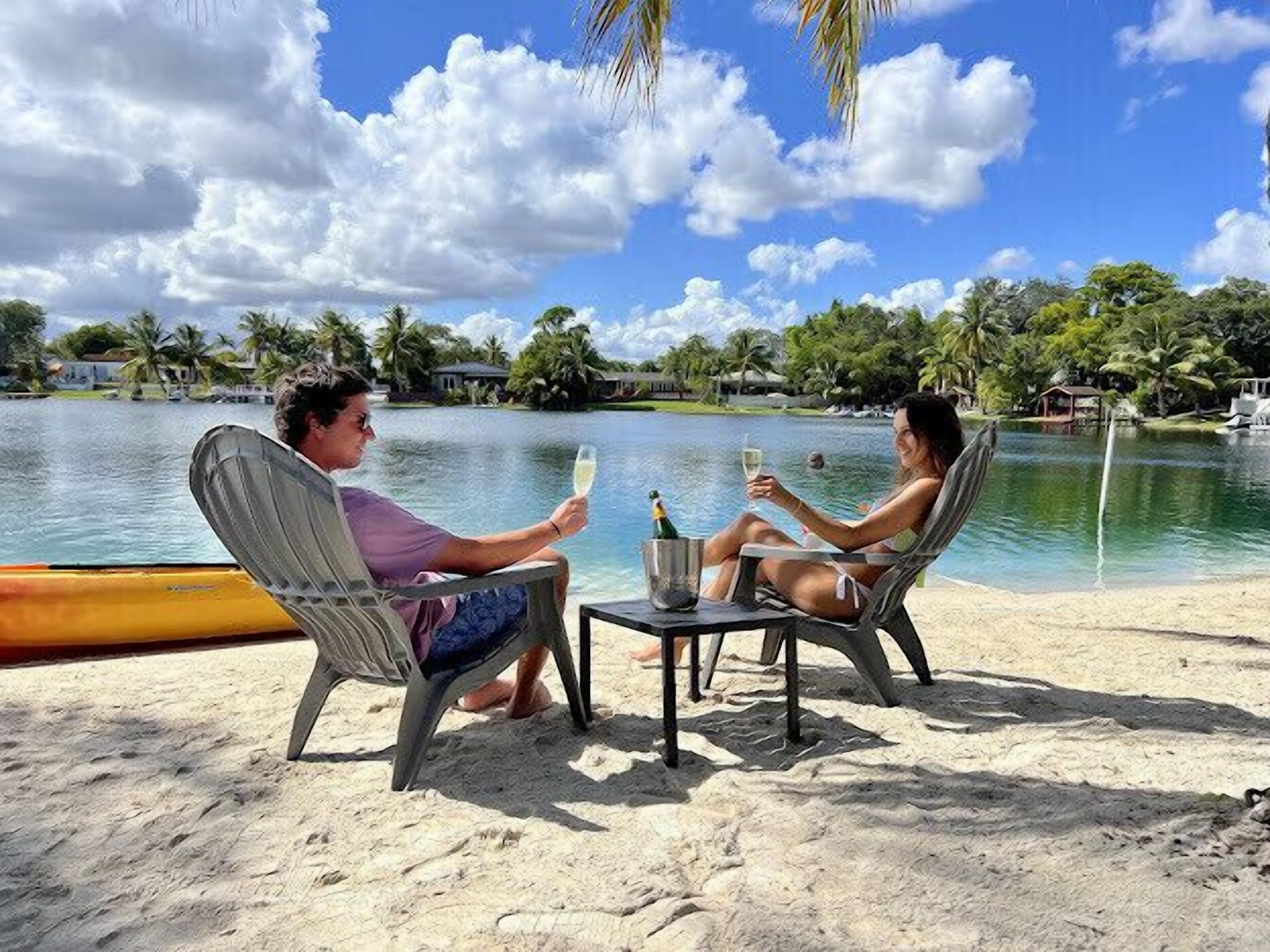 Dos personas sentadas en sillas de playa junto a una piscina, con vista al agua y palmeras.