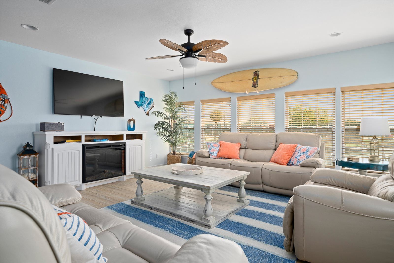 A living room with a flat-screen TV, a ceiling fan, a surfboard decoration, a fireplace, and a large window with blinds.
