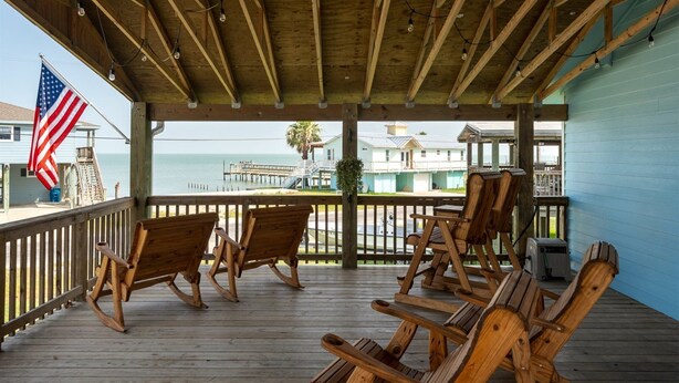 A wooden deck with rocking chairs, an American flag, and a view of a pier and buildings across the water. - Hook, Wine & Sinker by Miss Kitty’s (Rockport)